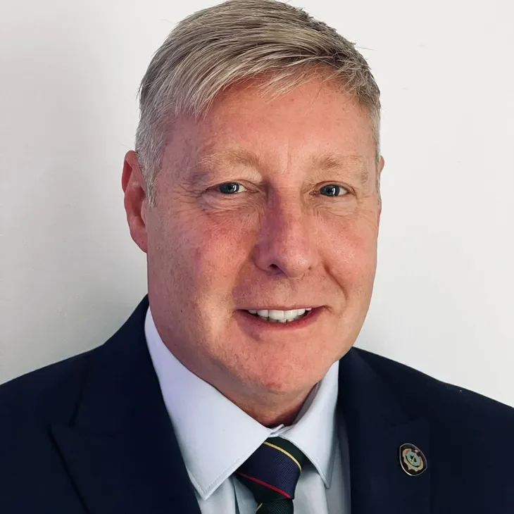 A man with short grey hair, smiling, wearing a dark suit and a tie.