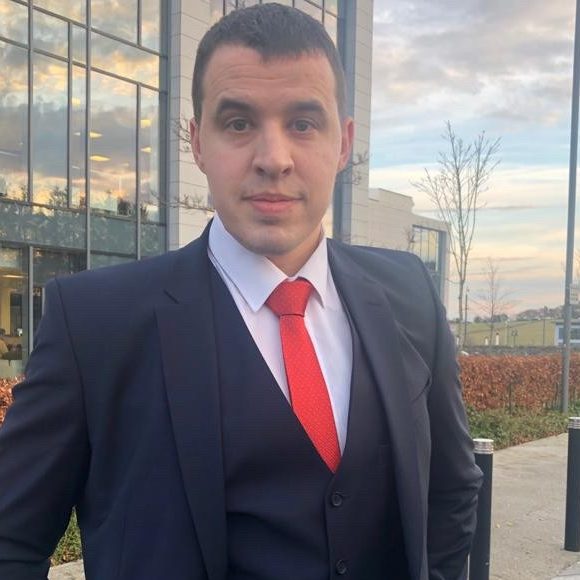 Man in a suit with a red tie, standing outside a modern building during the day.